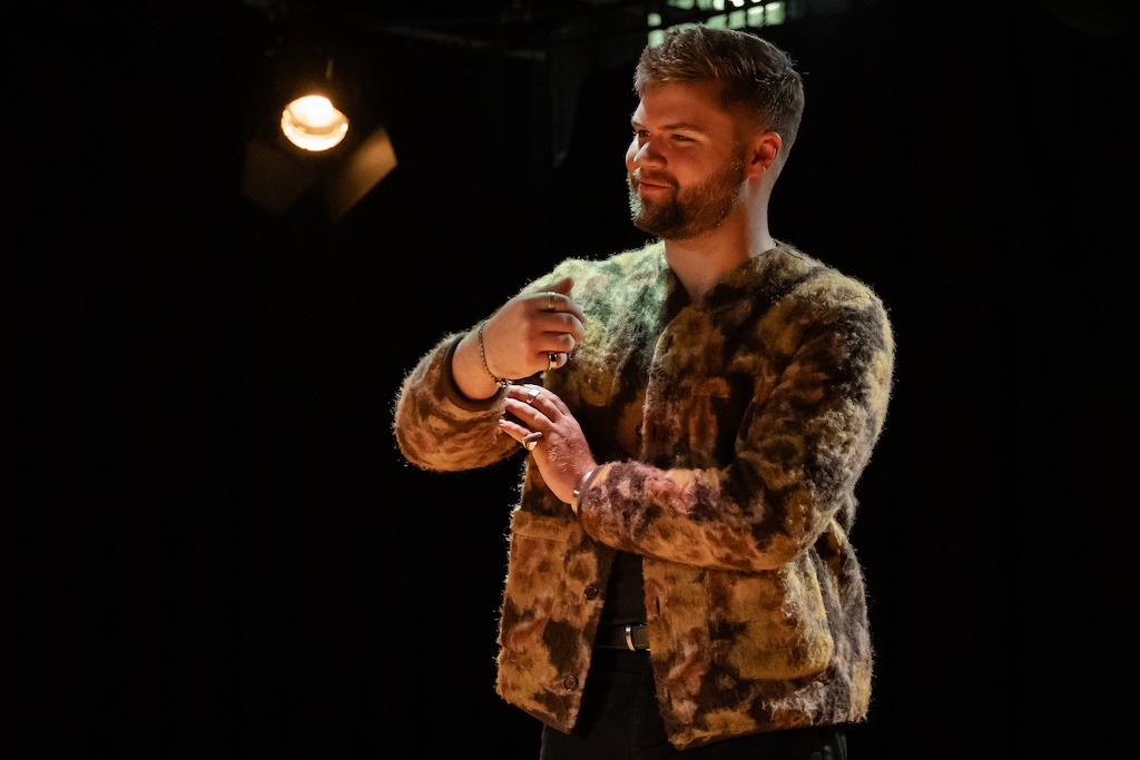 An actor wearing a patterned fur coat stands on stage in mid-performance during the final presentation of The Winter's Tale.